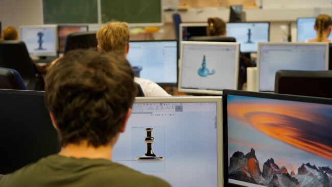 Ein junger Mann sitzt gemeinsam mit anderen Studierenden im Hörsaal einer Hochschule. Vor ihm steht ein Notebook.