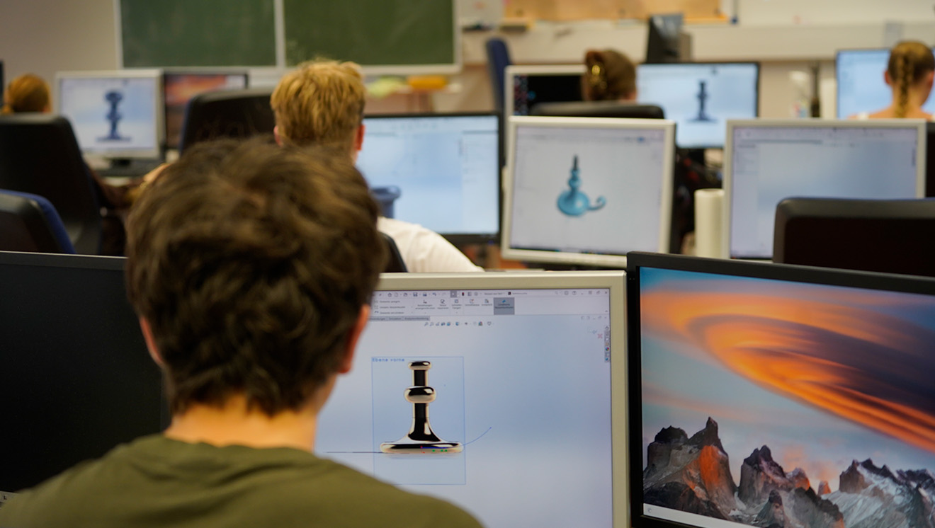 Ein junger Mann sitzt gemeinsam mit anderen Studierenden im Hörsaal einer Hochschule. Vor ihm steht ein Notebook.
